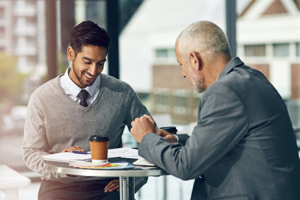 A young professional looking at his notepad while speaking with an older colleague across a desk in an outdoor setting.